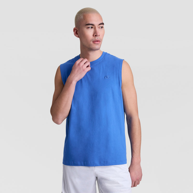 A person with short, light blonde hair wears a blue Champion Muscle Tank, C Logo, and white shorts against a plain light gray background, looking slightly to the side with one hand touching their collar.