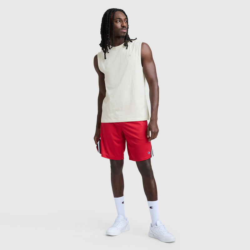 A man with long hair stands against a plain light background, wearing a beige Champion C Logo Muscle Tank, red athletic shorts, white crew socks, and white sneakers. He holds a phone in his right hand and looks slightly to the left.