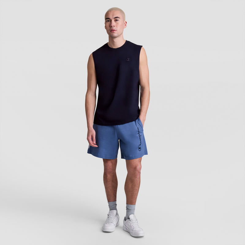 A man with a shaved head wears a black Champion Muscle Tank with the C Logo, blue shorts, gray socks, and white sneakers, standing against a plain light gray background.