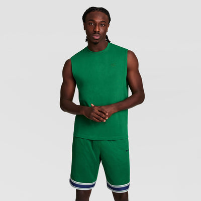 A man stands against a plain background, facing forward with hands together, wearing a green Champion Muscle Tank with C Logo and matching athletic shorts featuring white and dark blue stripes at the hem.