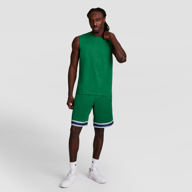A man stands against a plain light background wearing the Champion Muscle Tank, C Logo, with matching green shorts featuring white and navy striped trim, white socks, and white sneakers. He touches his hair and looks at the camera.