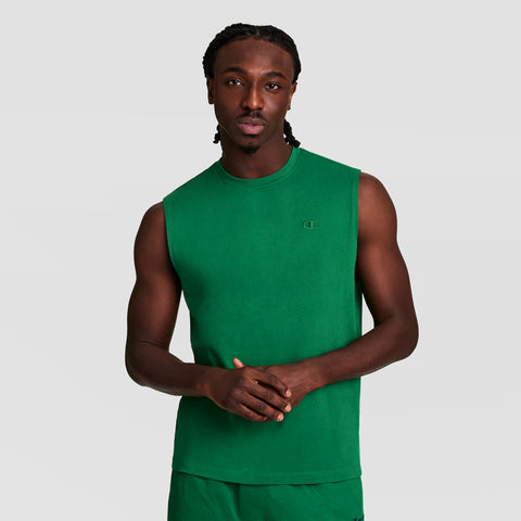 A man with braided hair wears a green Champion Muscle Tank featuring the C Logo and matching green shorts, standing against a plain light gray background with his hands folded in front of him.
