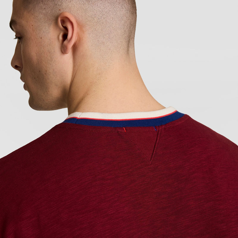 A person with short hair is seen from behind in a plain light-colored setting, wearing the Champion Modern Retro Washed Crew Sweatshirt, C Logo, featuring a maroon finish and a white, blue, and red striped collar.