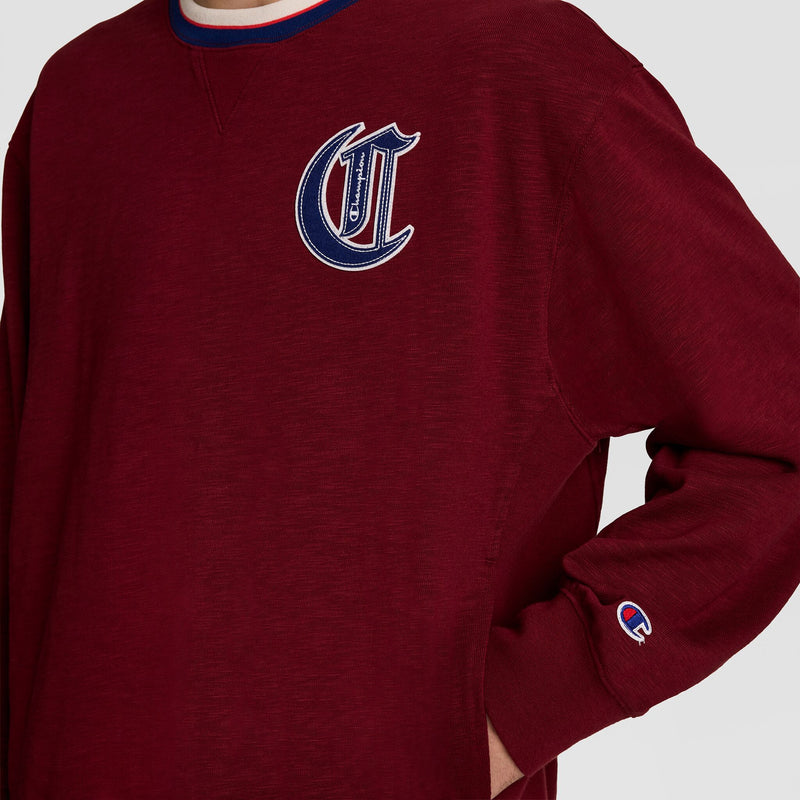 A person wearing the Champion Modern Retro Washed Crew Sweatshirt with a maroon, vintage streetwear look, large embroidered C logo on the chest, small C on the sleeve, and a ribbed collar featuring blue, white, and red stripes.
