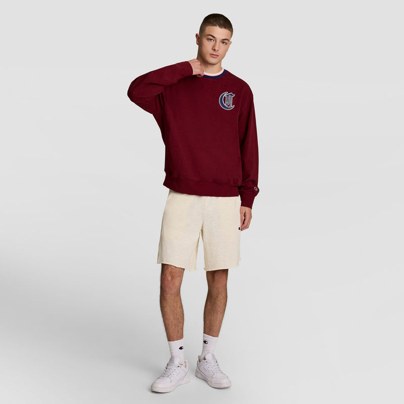 A young man stands against a plain background, wearing Champion’s Modern Retro Washed Crew Sweatshirt with a C logo in burgundy, off-white shorts, white socks, and sneakers. One hand rests on his neck.