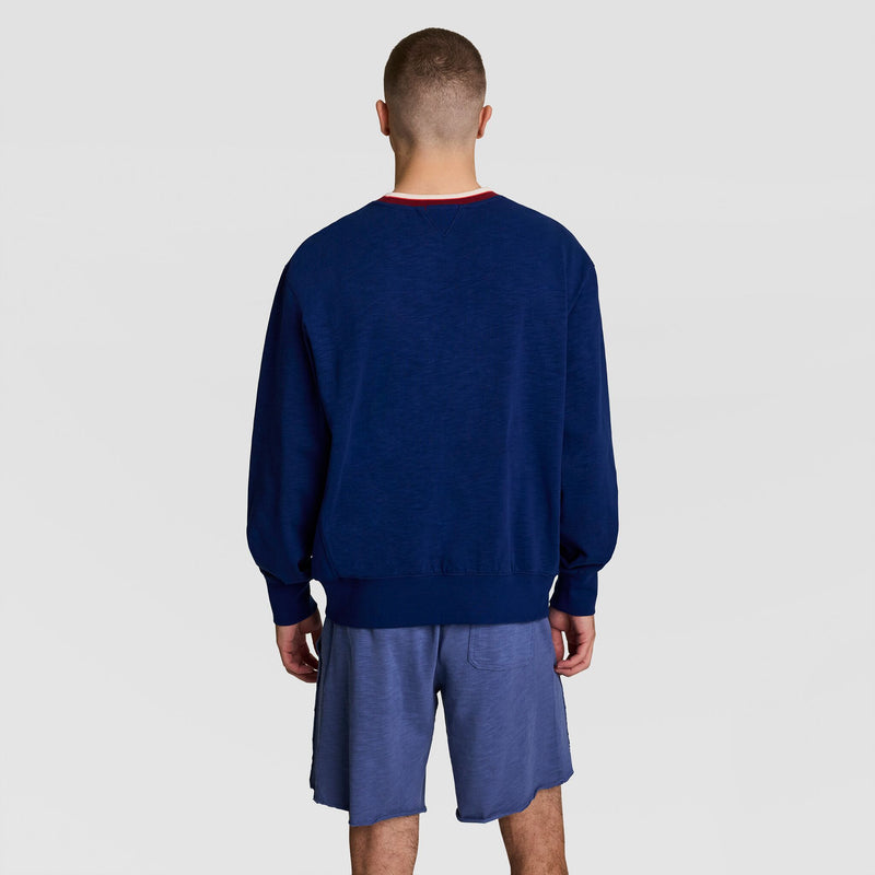 A person faces away from the camera, wearing a Champion Modern Retro Washed Crew Sweatshirt with C Logo in navy and blue athletic shorts against a plain light background, capturing a vintage streetwear look.