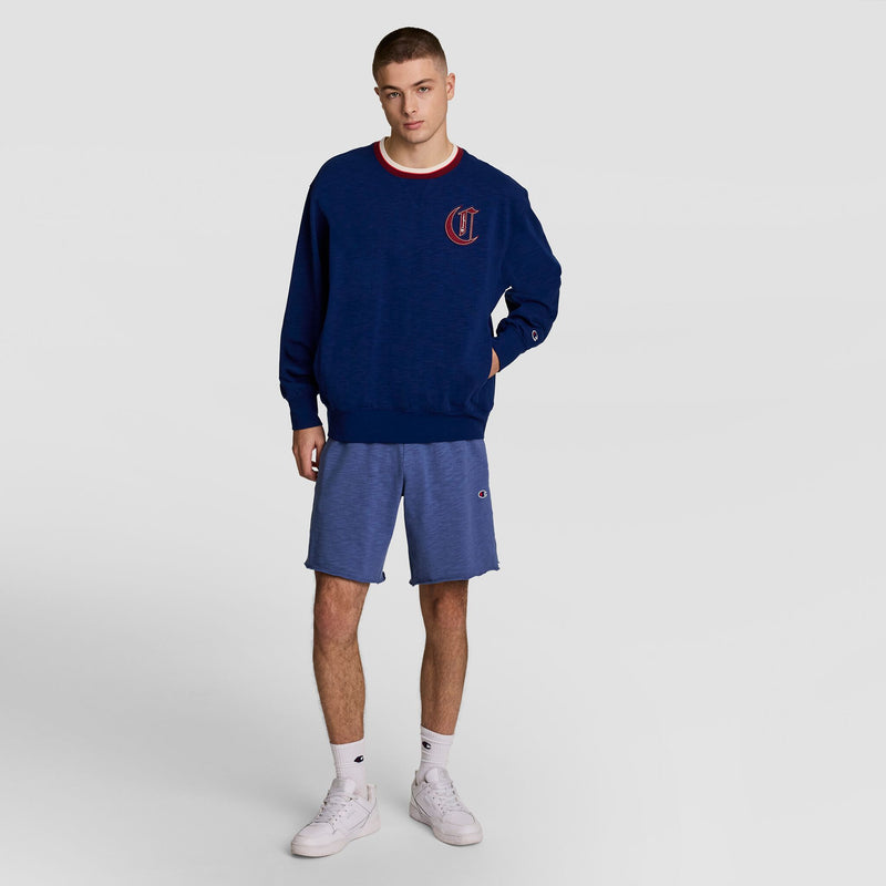 A young man stands facing forward in a Champion Modern Retro Washed Crew Sweatshirt with a red and white C logo, blue athletic shorts, white socks, and sneakers against a plain light gray background.