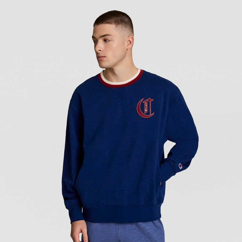 A young man with short brown hair wears the Champion Modern Retro Washed Crew Sweatshirt featuring a red C logo and a red and white collar, standing against a plain light gray background.