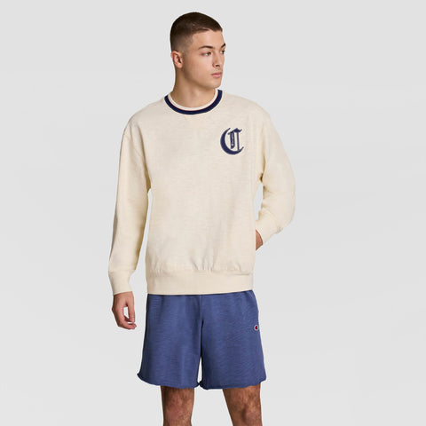 A young man in a Champion Modern Retro Washed Crew Sweatshirt with a navy “C” logo and blue shorts stands against a light gray background, capturing effortless streetwear style.