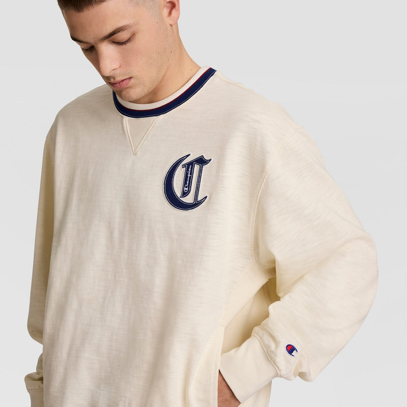 A young man wearing the Champion Modern Retro Washed Crew Sweatshirt featuring a cream color, navy “C” patch on the left chest, and Champion logo on the sleeve stands with one hand in his pocket, looking down.