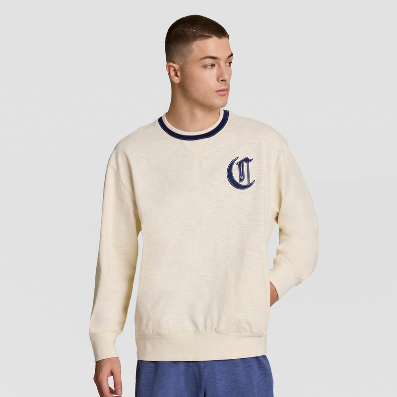 A young man wears the Champion Modern Retro Washed Crew Sweatshirt with a dark blue C logo, blue collar trim, and blue shorts, standing against a plain light background and looking to his left.