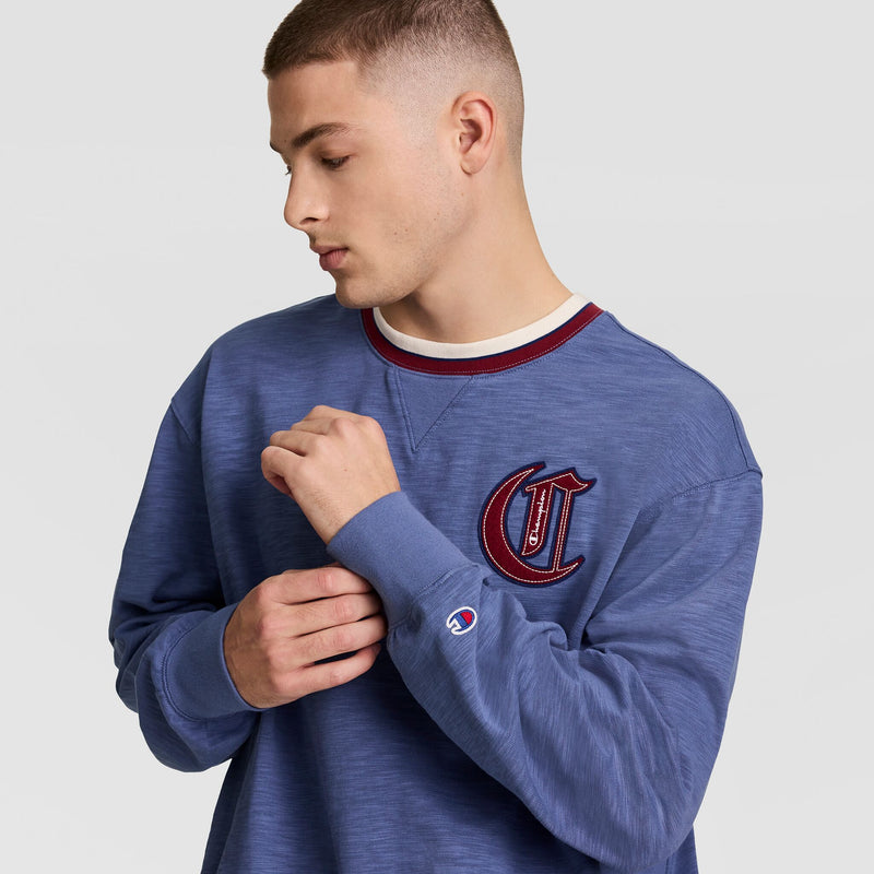 A young man with short hair wears the Champion Modern Retro Washed Crew Sweatshirt featuring a red and white C logo patch on the chest and a small sleeve logo, adjusting his sleeve against a plain gray background.