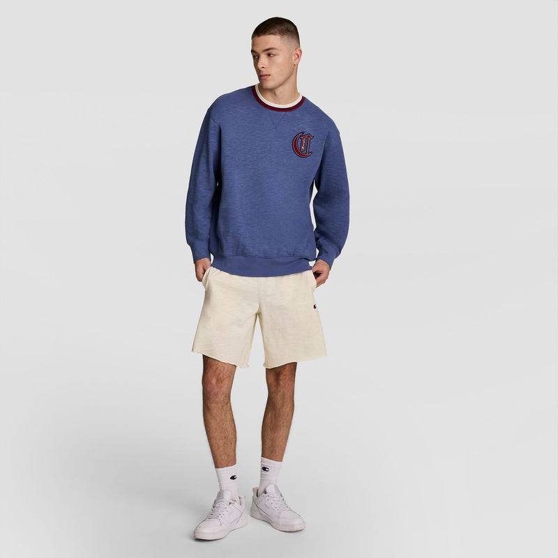 A young man stands against a plain background, wearing a Champion Modern Retro Washed Crew Sweatshirt with C Logo, cream shorts, white crew socks, and white sneakers. He looks to the side with hands in pockets, channeling modern streetwear style.