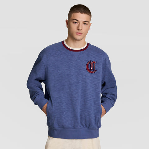 A young man with short hair models the Champion Modern Retro Washed Crew Sweatshirt, featuring a C logo on the chest, red and white collar, and blue heavy slub cotton, standing hands-in-pockets against a plain light background.
