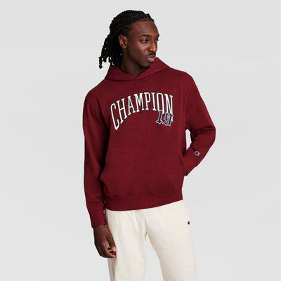 A person wears a maroon Champion Modern Retro Washed Hoodie with a C Logo, paired with cream-colored pants, standing against a plain light gray background.