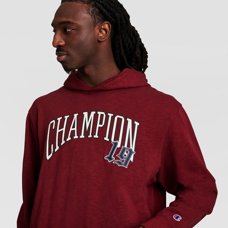 A person with long braided hair wears a maroon Champion Modern Retro Washed Hoodie featuring the C Logo, looking to the side against a plain background.