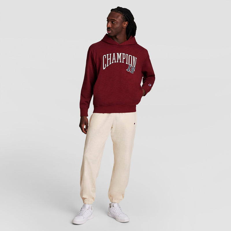 A man in a maroon Champion Modern Retro Washed Hoodie with C Logo, off-white jogger pants, and white sneakers stands against a plain light gray background, looking to his left with one hand behind his back.