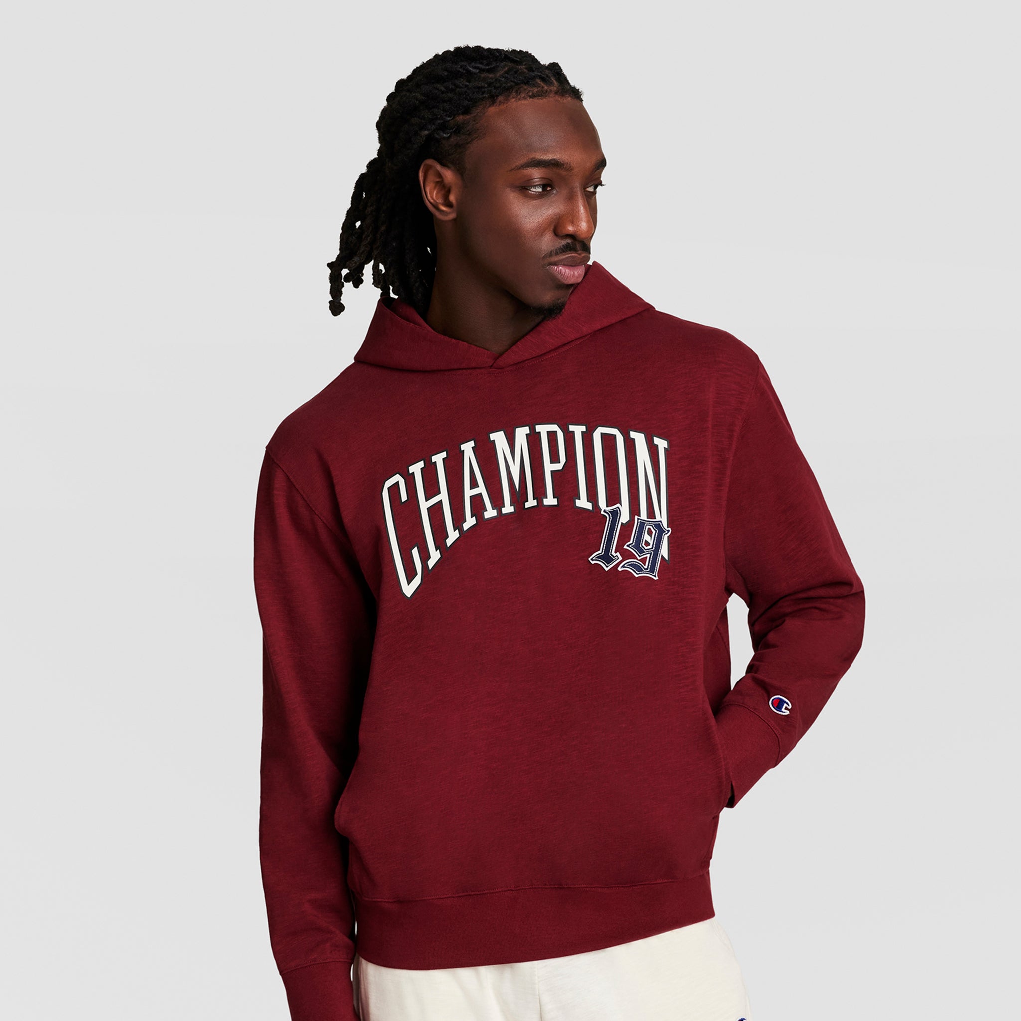 A man in a maroon Champion Modern Retro Washed Hoodie with a white C logo and "19" patch stands against a plain light background, looking left with one hand in his pocket, highlighting vintage athletic style.