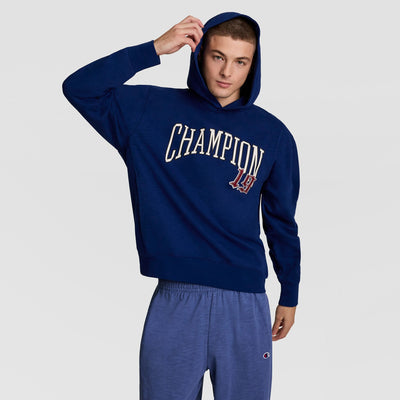 A young man in a blue Champion Modern Retro Washed Hoodie with a C logo and matching sweatpants adjusts his hood while posing against a plain light gray background.