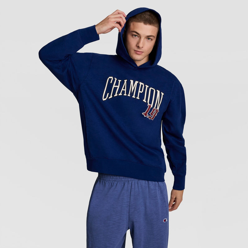 A young man in a blue Champion Modern Retro Washed Hoodie with a C logo and matching sweatpants adjusts his hood while posing against a plain light gray background.