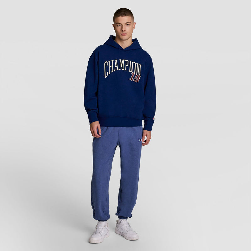 A young man stands against a plain background, wearing a navy blue Champion Modern Retro Washed Hoodie with the C logo, blue jogger pants, and white sneakers.