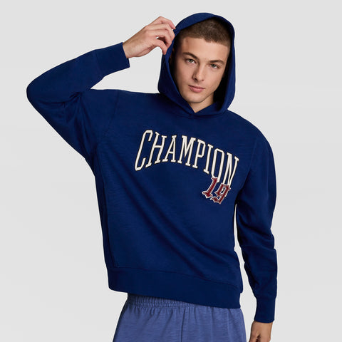 A young man in a blue Champion Modern Retro Washed Hoodie featuring the C logo and blue shorts stands against a plain background, lifting the hood with one hand while looking at the camera.