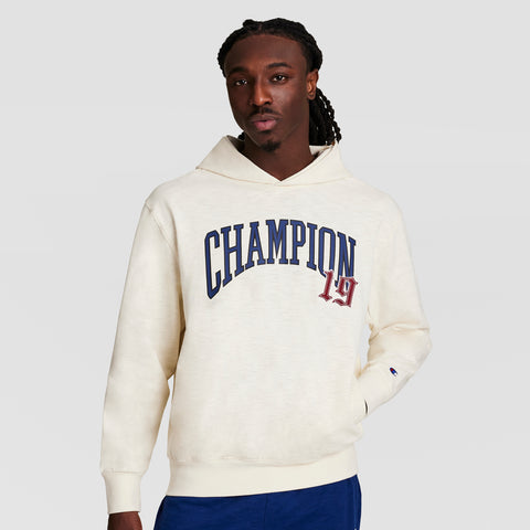 A man with long braids wears the Champion Modern Retro Washed Hoodie, C Logo, in off-white with bold blue and red CHAMPION 19 lettering. The plain light background highlights this stylish retro-inspired hoodie.