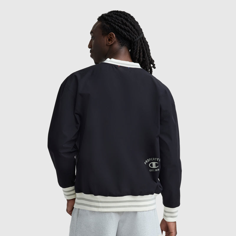 A man with long, braided hair wears a black Champion Gridiron V-Neck Pullover with a small C logo near the bottom hem and white trim, paired with light gray shorts, standing with his back to the camera. The pullover is water-resistant.