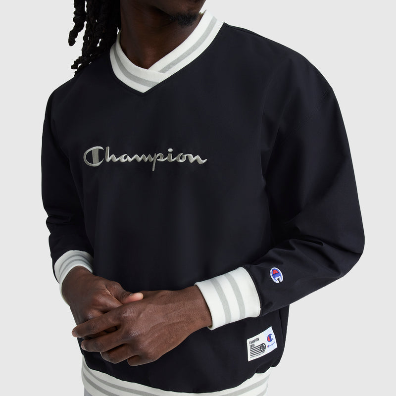 A person wears a black Champion Gridiron V-Neck Pullover with white striped cuffs and collar. Water-resistant fabric and embroidered C logo add sporty style. The person's face is partially out of frame.