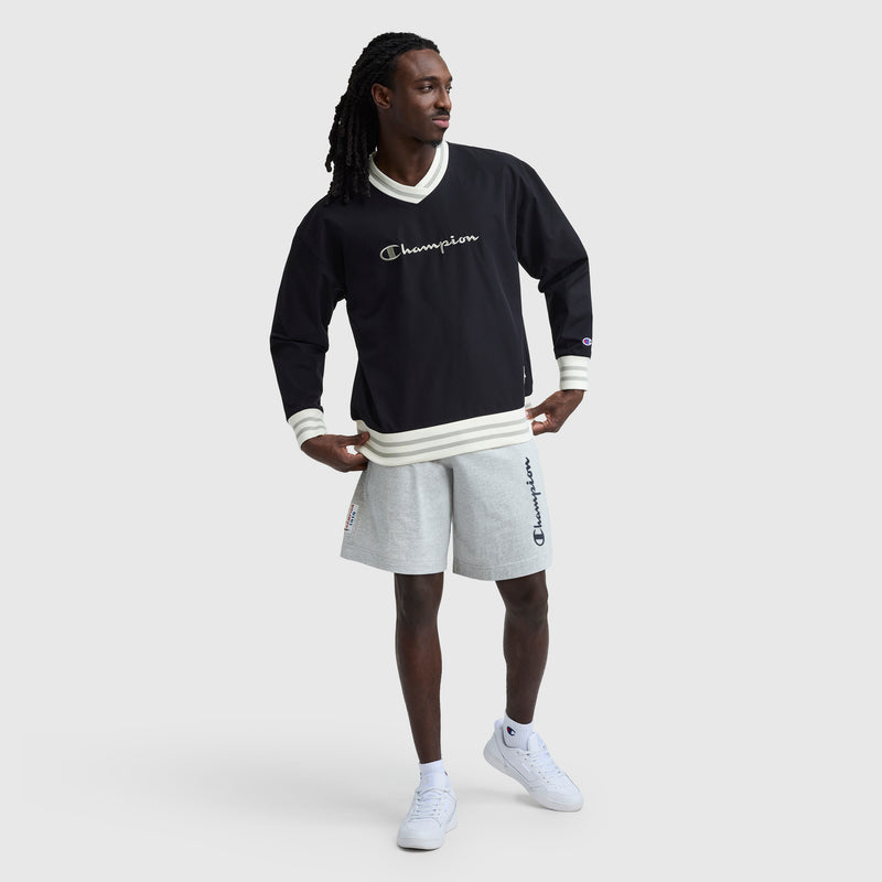 A man in a black Champion Gridiron V-Neck Pullover with C Logo, white trim, light gray Champion shorts, and white sneakers stands against a plain light background, looking to the side with his hands on his hips.