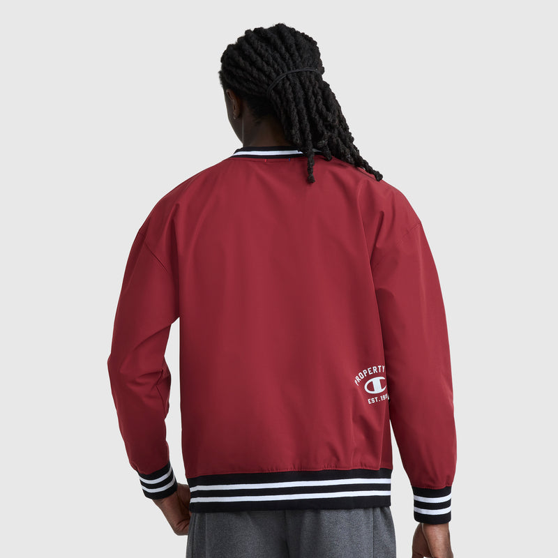 A person with long, braided hair faces away wearing a red Champion Gridiron V-Neck Pullover. The water-resistant jacket has black and white striped cuffs and waistband, plus a small white C logo near the bottom hem.