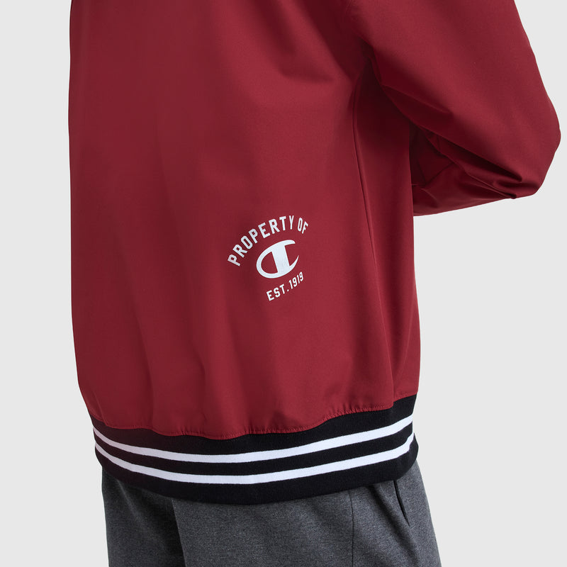 A person wearing a red Champion Gridiron V-Neck Pullover with a C logo, "PROPERTY OF C EST. 1919" printed in white on the back, gray pants, and a black and white striped waistband.