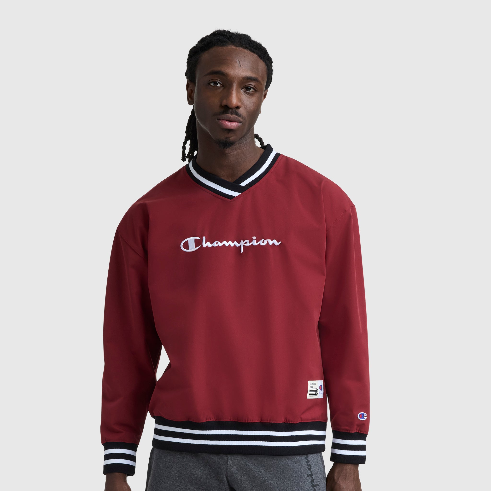 A man with long braids wears a maroon Champion Gridiron V-Neck Pullover featuring the C Logo and black-and-white striped cuffs and collar, standing against a plain light gray background.