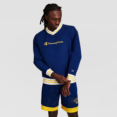 A man in a blue Champion Gridiron V-Neck Pullover with C Logo and yellow/white stripes, plus matching blue shorts with yellow trim, stands against a white background, looking down while adjusting his sleeve.