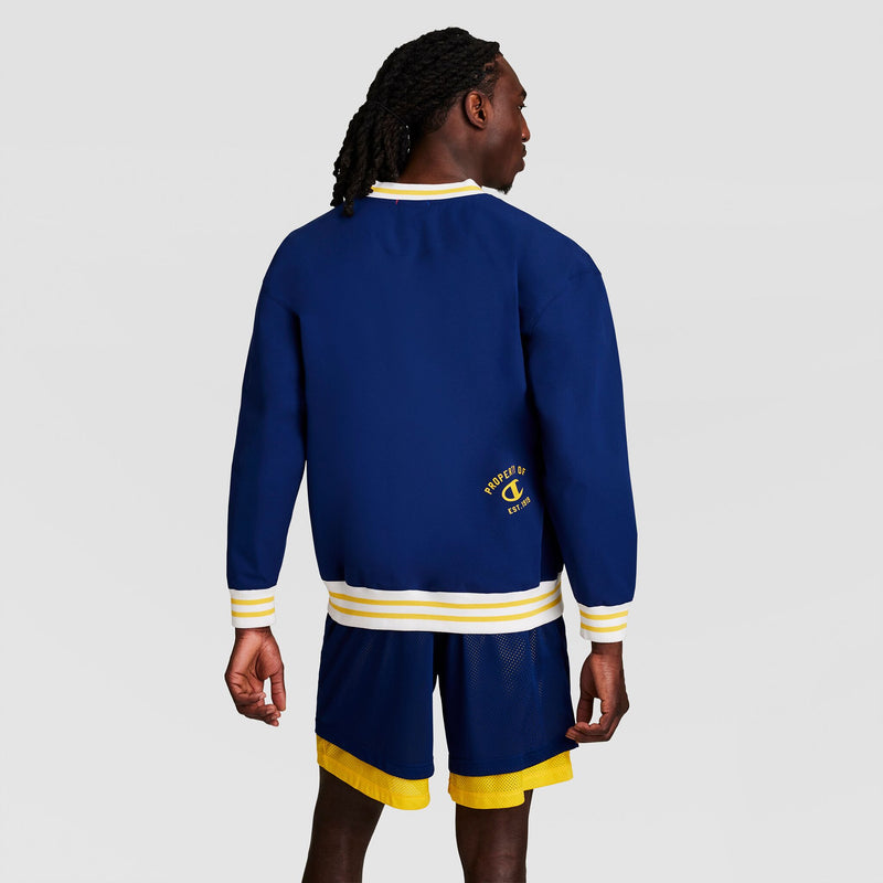 A person with long dark hair wears the Champion Gridiron V-Neck Pullover with C Logo in blue and yellow, standing with their back to the camera against a plain, light background.