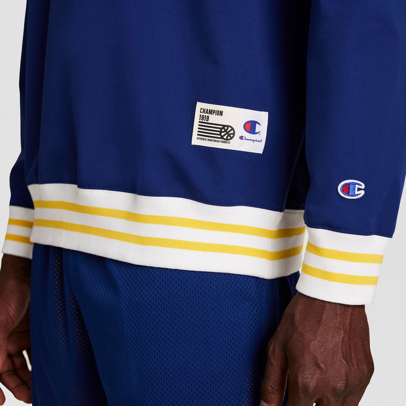 A person wears the Champion Gridiron V-Neck Pullover in blue, featuring white and yellow striped cuffs and hem, water-resistant fabric, a C logo on the sleeve, and a CHAMPION 1919 label above the hem.