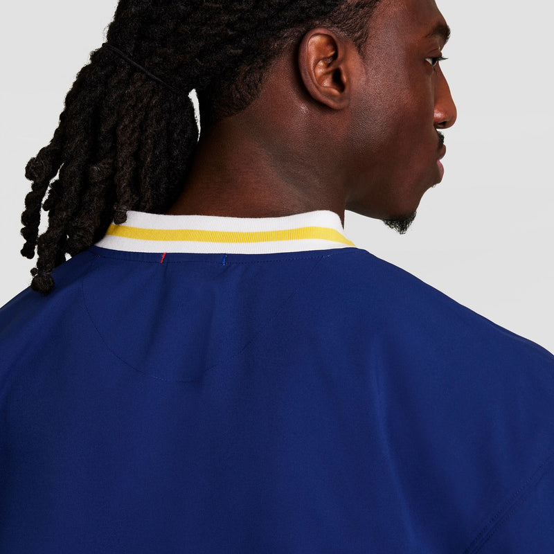 A person with long, dark, braided hair is shown from the back and side, wearing a blue Champion Gridiron V-Neck Pullover with a white and yellow collar. The light background is plain. The pullover features the C Logo.