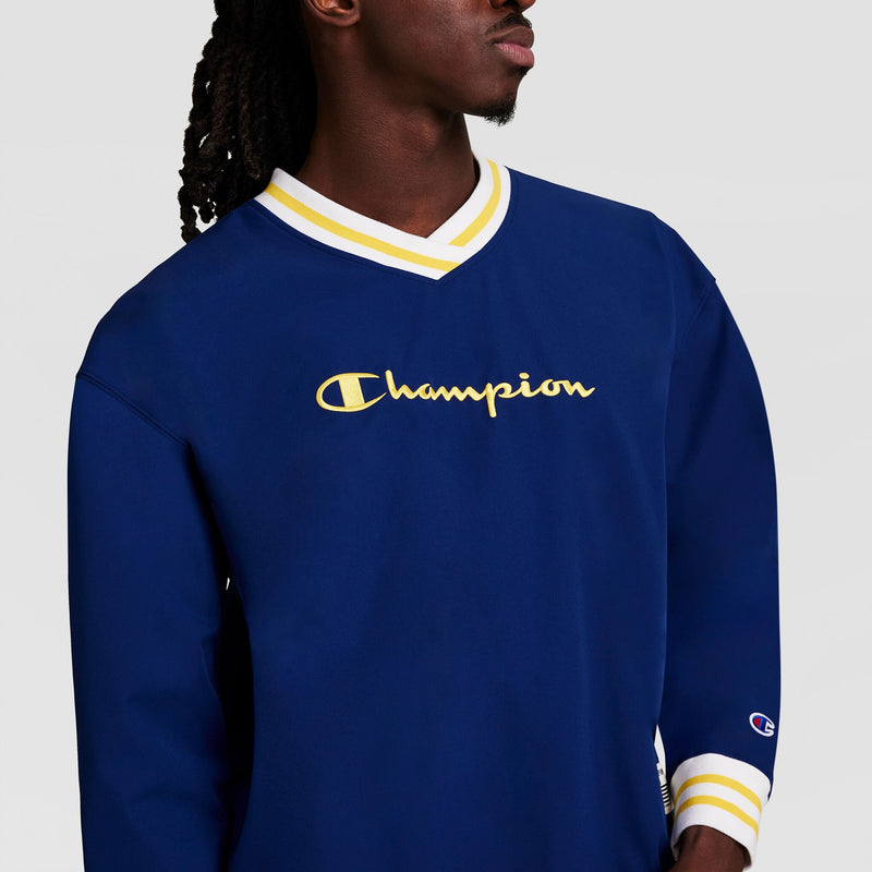 Someone wears a blue Champion Gridiron V-Neck Pullover with C Logo, featuring yellow and white trim on the collar and cuffs. "Champion" is embroidered in yellow across the chest. The background is plain and light-colored.