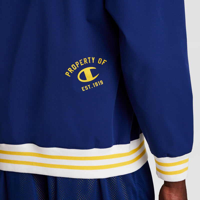 A person wears a blue, water-resistant Champion Gridiron V-Neck Pullover with white and yellow striped trim. "PROPERTY OF C EST. 1919" is printed in yellow on the back, with the Champion C logo underneath.