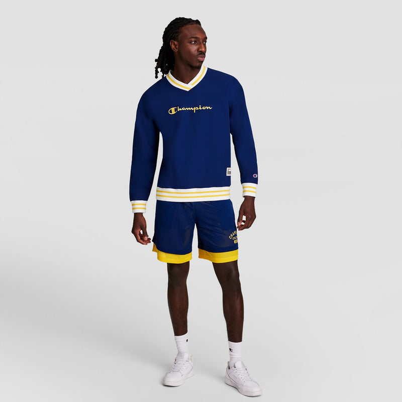 A man in a blue Champion Gridiron V-Neck Pullover with C Logo and matching blue shorts with yellow trim stands against a plain light background, looking to the side and wearing white sneakers.