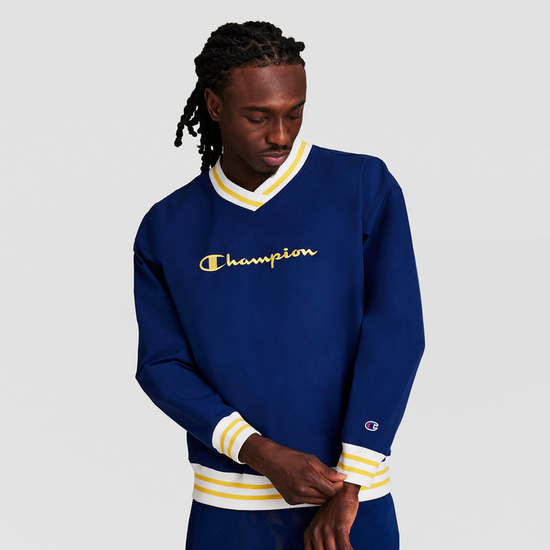 A man adjusts his sleeve while wearing a navy blue Champion Gridiron V-Neck Pullover with C Logo and yellow and white striped cuffs and collar, standing against a plain light background.