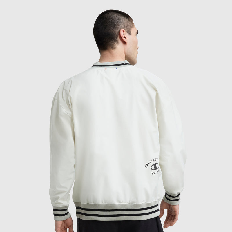A person in a white Champion Gridiron V-Neck Pullover with gray and black striped cuffs and collar stands with their back to the camera. The water-resistant fabric and C Logo are visible on the lower back of the pullover.