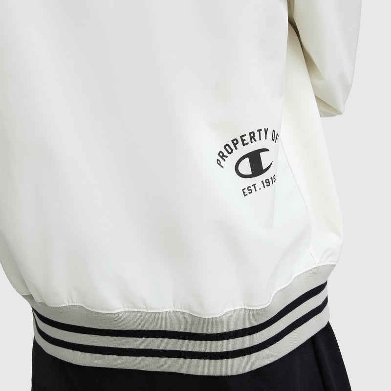 A close-up of the lower back of a white Champion Gridiron V-Neck Pullover featuring striped gray and black trim, water-resistant fabric, the Champion C logo, and "PROPERTY OF EST. 1919" printed in black text.