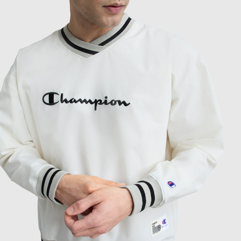 A person wears the Champion Gridiron V-Neck Pullover with the C logo, featuring black and gray striped cuffs and collar. The Champion branding is on the chest, with a small patch on the wrist. The face is partially visible.