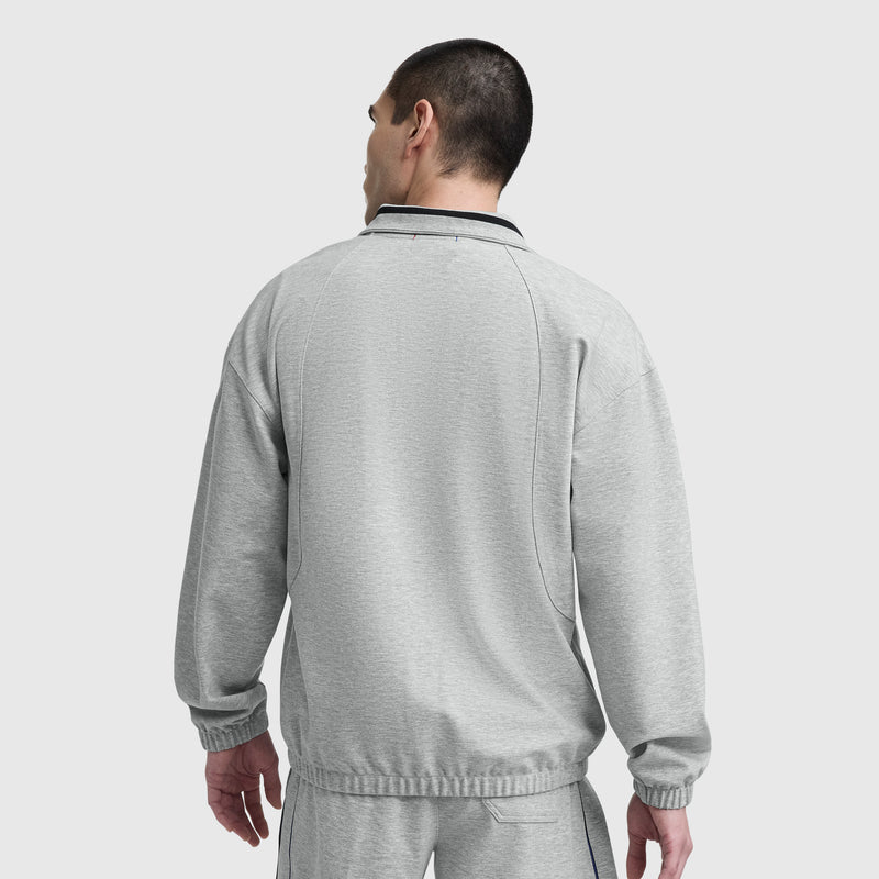 A man with short dark hair stands with his back to the camera, wearing Champion’s Gameday Full Zip Mock Neck and matching pants with black trim. An embroidered C Logo adds a vintage athletic touch against the plain light background.