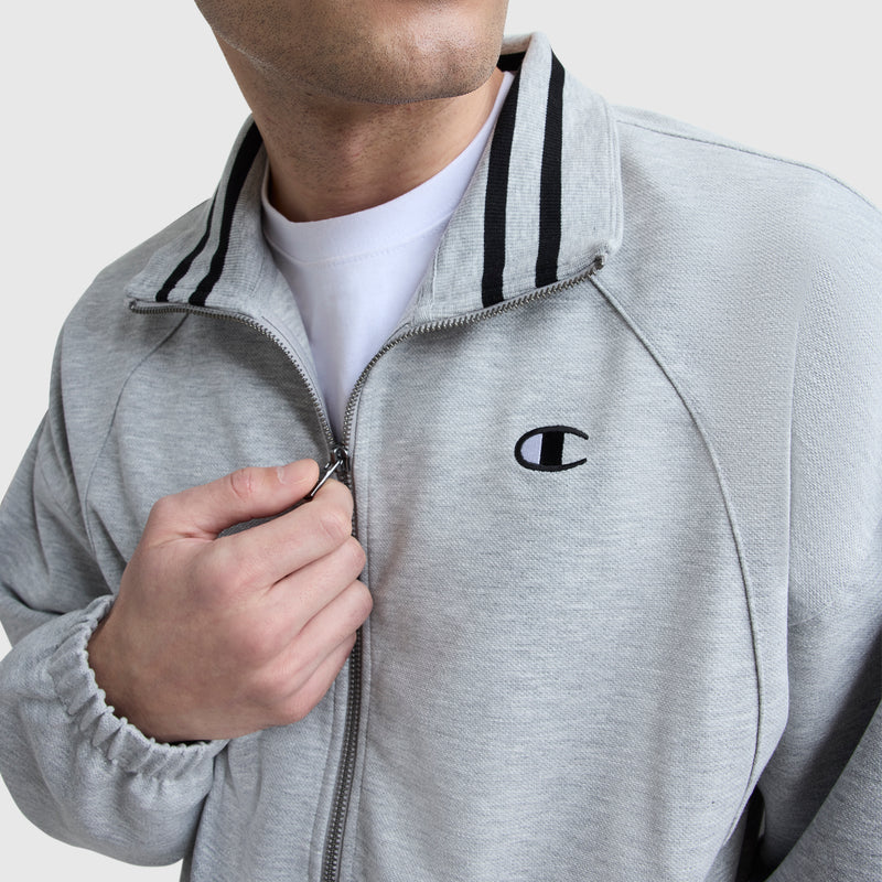 A person zips up a light gray Champion Gameday Full Zip Mock Neck jacket with black-striped collar, featuring an embroidered C logo on the chest and vintage athletic design, worn over a white T-shirt.