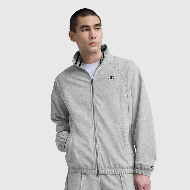 A man stands with one hand in his pocket against a plain light background, wearing a light gray Champion Gameday Full Zip Mock Neck jacket featuring an embroidered C logo and matching pants.