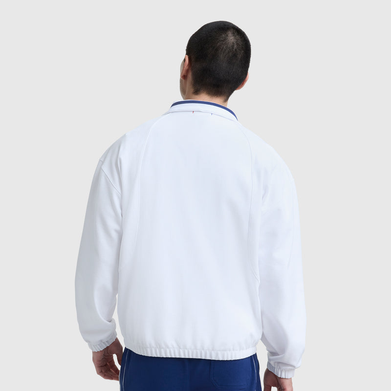 A person with short dark hair faces away from the camera, wearing the Champion Gameday Full Zip Mock Neck athletic jacket with C Logo in white and blue pants, evoking vintage sport style against a plain light gray background.