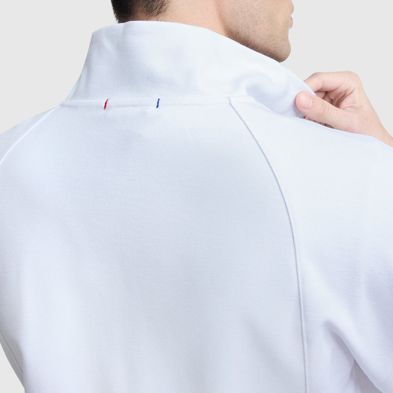 A person adjusts the collar of a Champion Gameday Full Zip Mock Neck jacket, featuring C Logo details with small red and blue stitches near the collar seam. The white athletic jacket is shown from the back against a plain, light background.