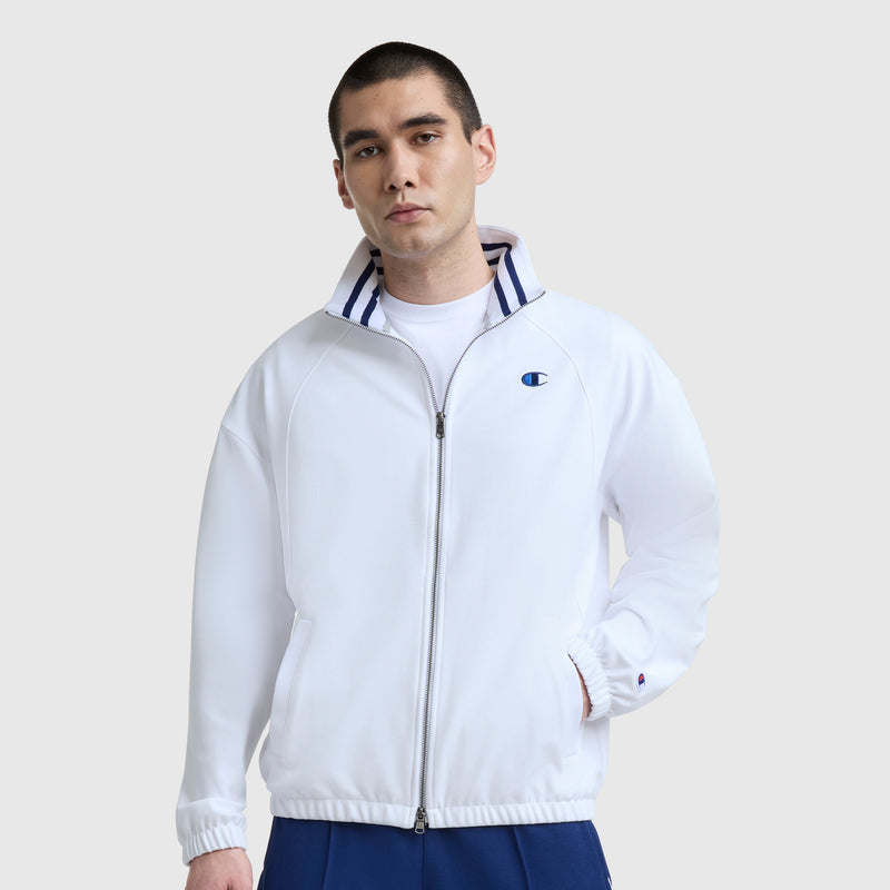 A man in a Champion Gameday Full Zip Mock Neck, C Logo jacket with blue collar stripes and blue pants stands against a plain light gray background, wearing a neutral expression.